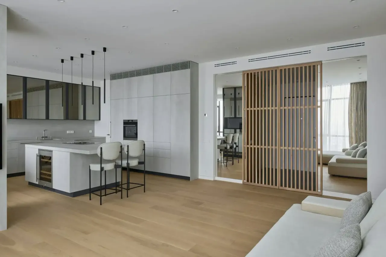 Open-plan kitchen with a white island and bar stools beside a living area behind a wood slat screen