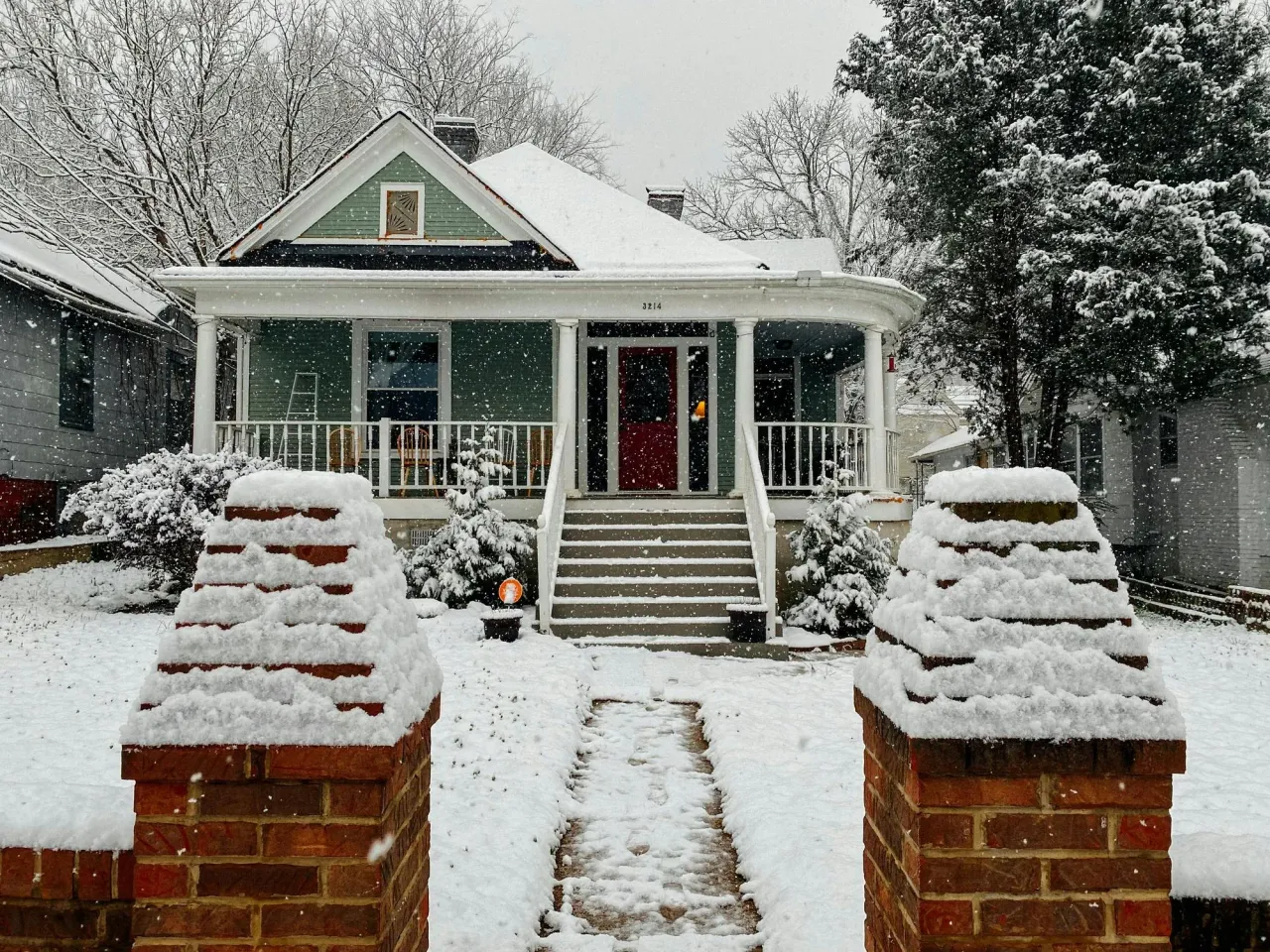 Older bungalow with a covered front porch and brick gate pillars in falling snow